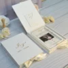 Ivory velvet invitation box with a gold foil monogram next to an open box with a clear acrylic wedding invitation over a bride and groom photo, a lid pocket of insert cards and ivory roses.